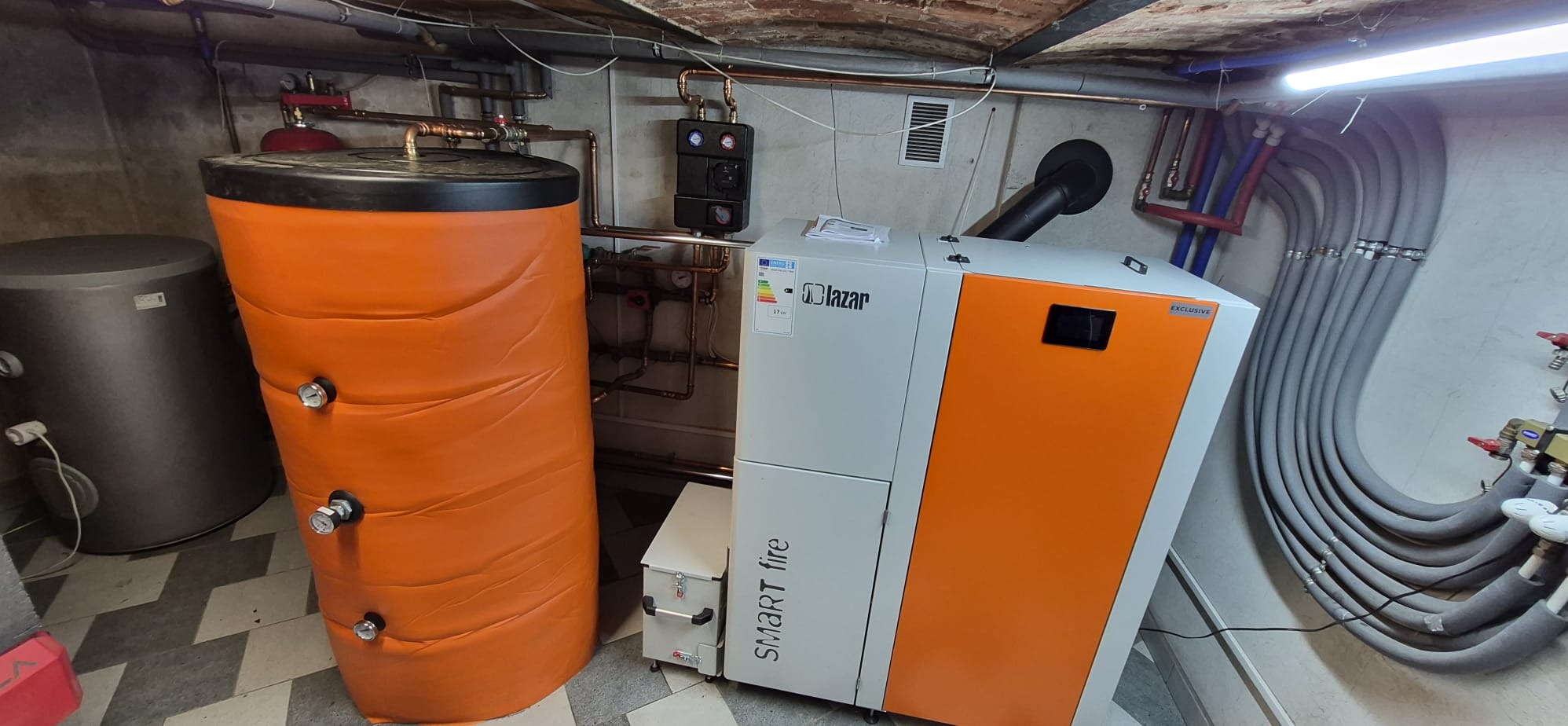 Kompleksowy montaż kotła na pellet Lazar Smart Fire z zasobnikiem i zbiornikiem buforowym. Widoczne rury instalacji grzewczej i osprzęt w kotłowni.