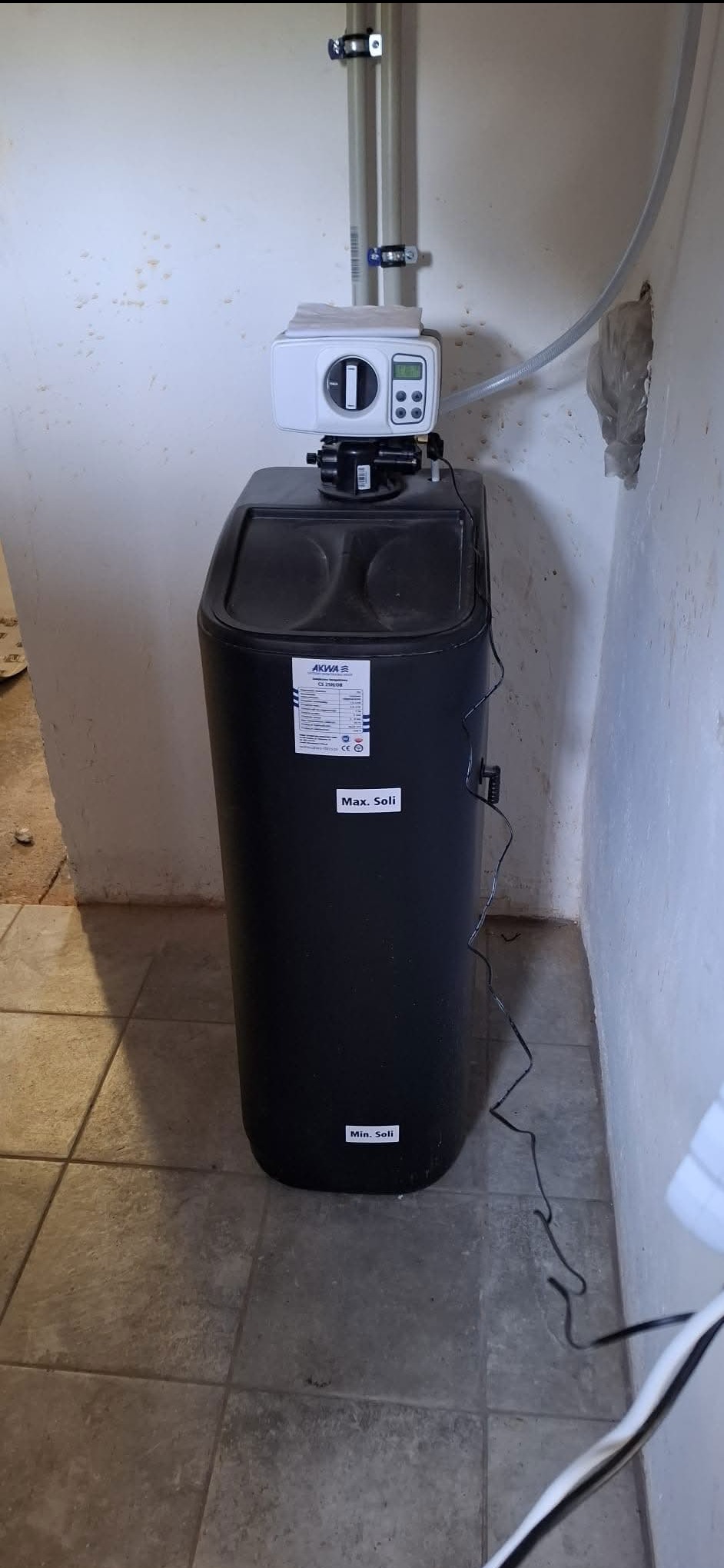 Czarna stacja uzdatniania wody Akwa Soli z białym sterownikiem, zainstalowana w pomieszczeniu z szarą podłogą i ścianami z widocznymi śladami. Widoczne rury i przewody.