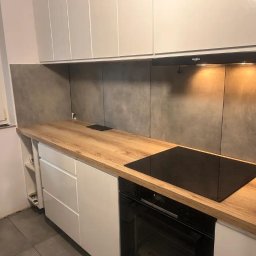 WARM HOME Adrian Laskowski - Nowoczesna kuchnia z drewnianym blatem, białymi szafkami i szarymi płytkami. Widoczna płyta indukcyjna i okap. Minimalistyczny design z elementami wykończenia.