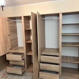 WARM HOME Adrian Laskowski - Jasne, drewnopodobne szafy z otwartymi drzwiami i wysuniętymi szufladami, ukazujące wnętrze z półkami i drążkami na ubrania. Widok na podłogę z paneli.