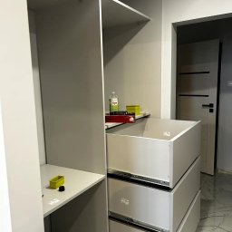 WARM HOME Adrian Laskowski - Montaż szafy wnękowej: widoczne szuflady na prowadnicach, półki i akcesoria montażowe. W tle marmurowa podłoga i drzwi. Ujęcie z bliska, podkreślające detale.