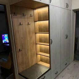 WARM HOME Adrian Laskowski - Funkcjonalna szafa w przedpokoju z siedziskiem, wieszakami i podświetlanymi półkami. Fronty w odcieniach bieli z widoczną strukturą drewna, czarne uchwyty.