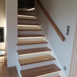 WARM HOME Adrian Laskowski - Nowoczesne schody drewniane z subtelnym podświetleniem LED, białe ściany, drewniana poręcz. Minimalistyczny design z akcentem na oświetlenie.