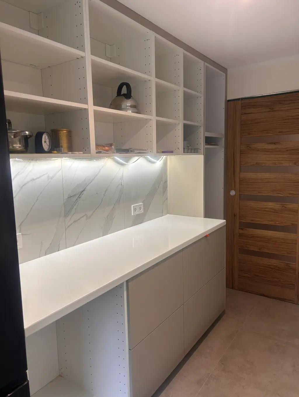 Jasna kuchnia z białymi szafkami i blatem, marmurowym backsplashiem oraz drewnianymi drzwiami przesuwnymi. Na półkach czajnik i dekoracje. Minimalistyczny design.