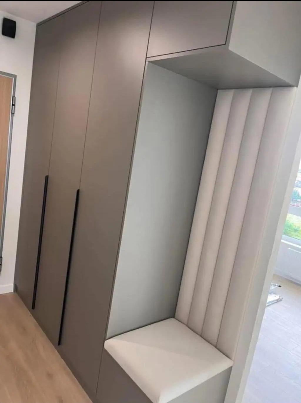 Szara szafa wnękowa z siedziskiem obitym jasną tkaniną, wkomponowana w przedpokój. Minimalistyczne, długie uchwyty. Podłoga z paneli imitujących drewno.