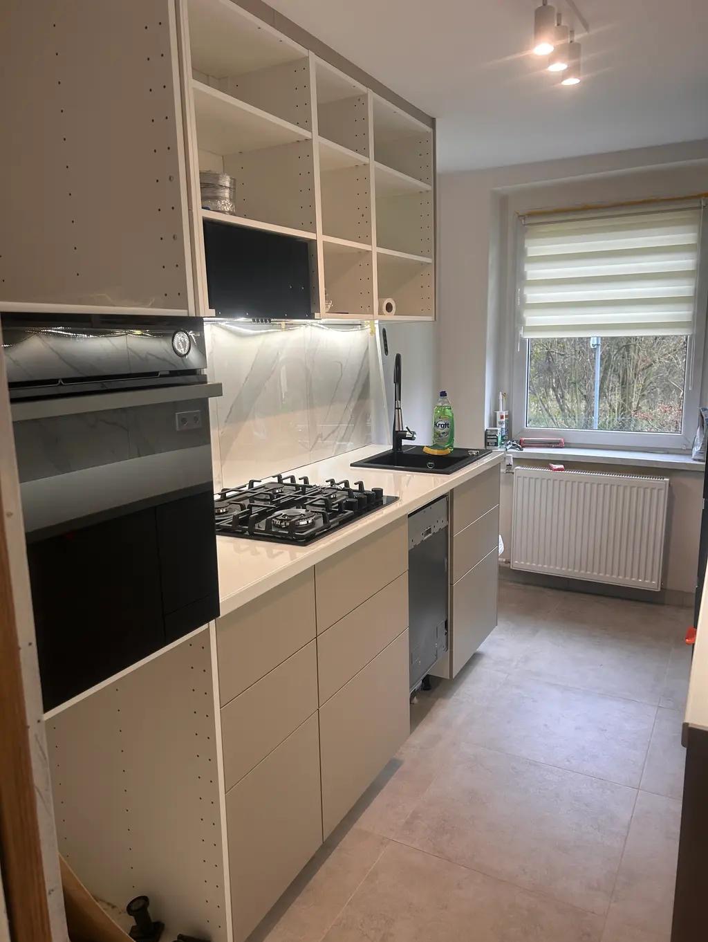 Jasna kuchnia z białymi szafkami bez frontów, marmurowym backsplashiem i czarnym zlewem. Widoczna płyta gazowa i piekarnik. Minimalistyczny design, neutralna kolorystyka.