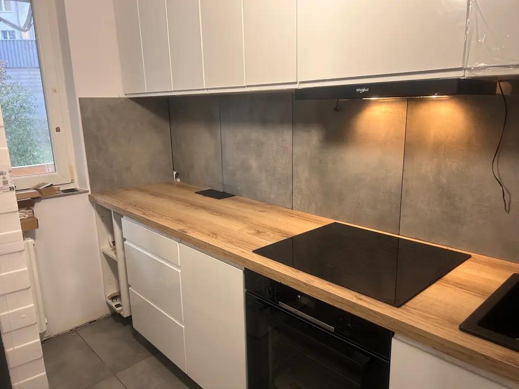 Nowoczesna kuchnia z drewnianym blatem, białymi szafkami i szarymi płytkami. Widoczna płyta indukcyjna i okap. Minimalistyczny design z elementami wykończenia.