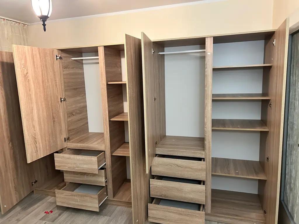 Jasne, drewnopodobne szafy z otwartymi drzwiami i wysuniętymi szufladami, ukazujące wnętrze z półkami i drążkami na ubrania. Widok na podłogę z paneli.