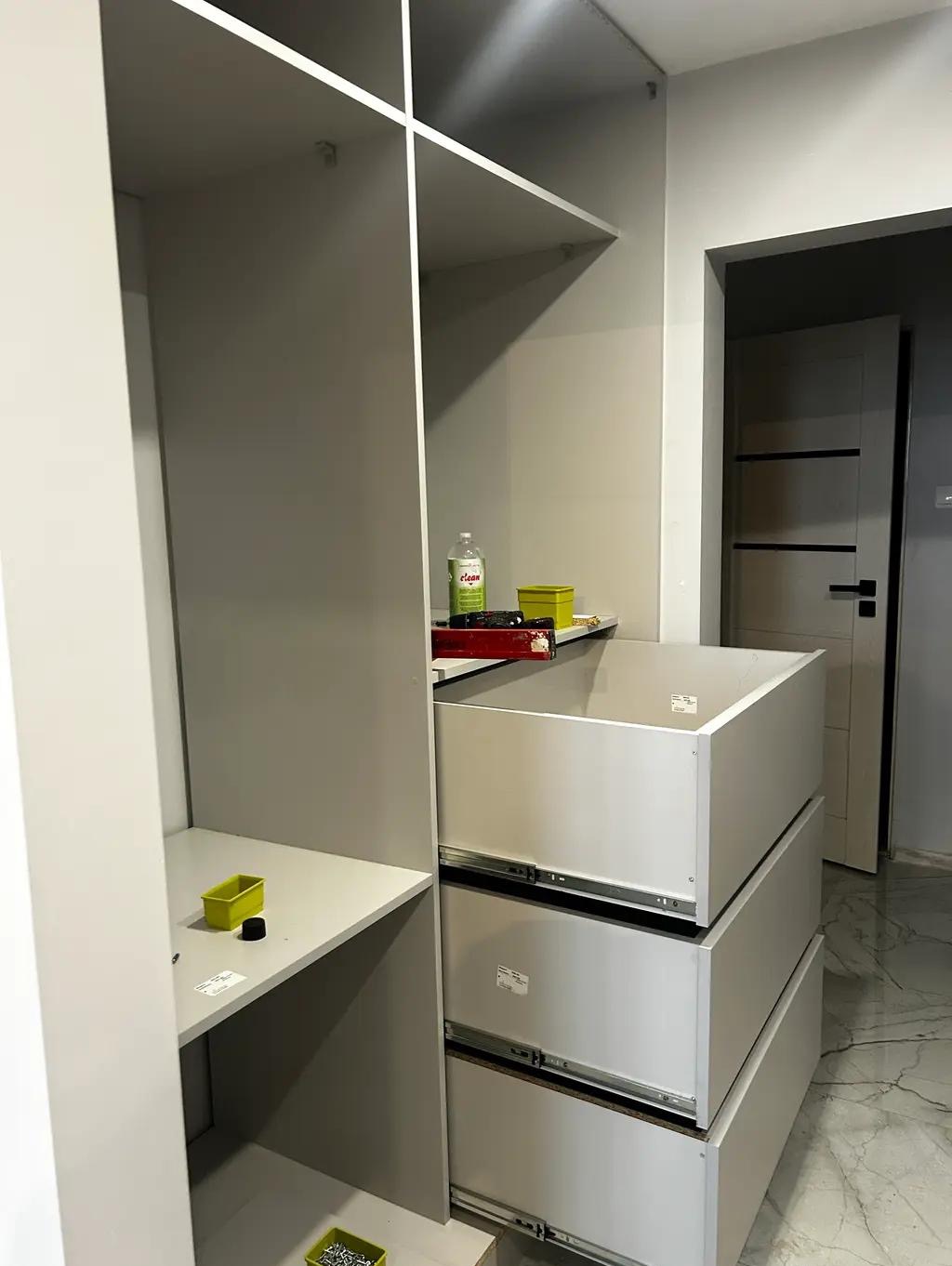 Montaż szafy wnękowej: widoczne szuflady na prowadnicach, półki i akcesoria montażowe. W tle marmurowa podłoga i drzwi. Ujęcie z bliska, podkreślające detale.