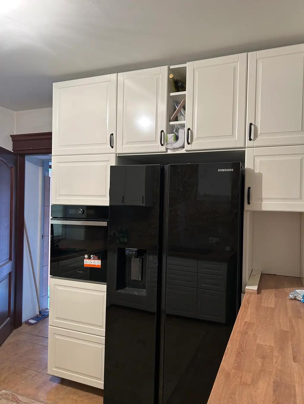 Biała zabudowa kuchenna z czarną lodówką Samsung, widok na szafki górne z otwartą półką na wino i piekarnik w zabudowie. Ujęcie z perspektywy podłogi.