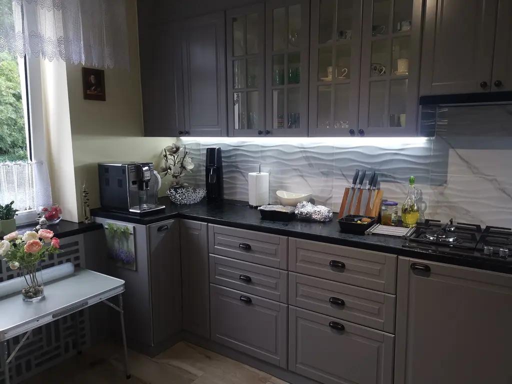 Szara kuchnia z frezowanymi frontami, czarnym blatem i falistym backsplashiem. Na blacie ekspres do kawy, akcesoria kuchenne i płyta gazowa. Widok na okno z firanką.