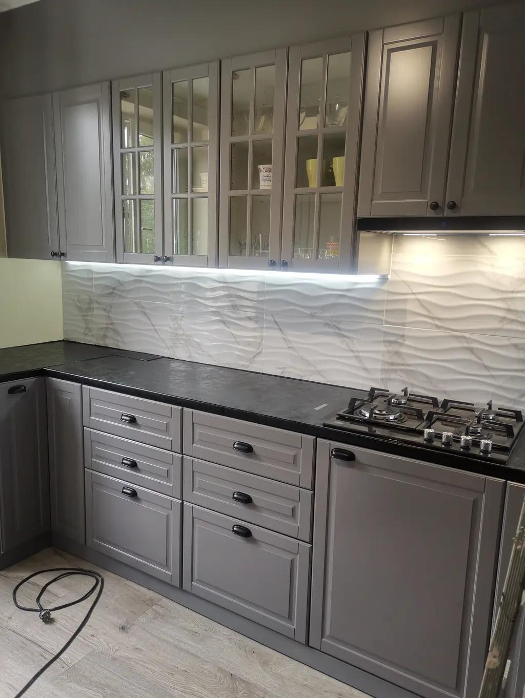 Szara kuchnia z frezowanymi frontami, czarnym blatem i płytkami 3D. Górne szafki przeszklone, oświetlenie LED podszafkowe. Nowoczesny design, widok na fragment podłogi.