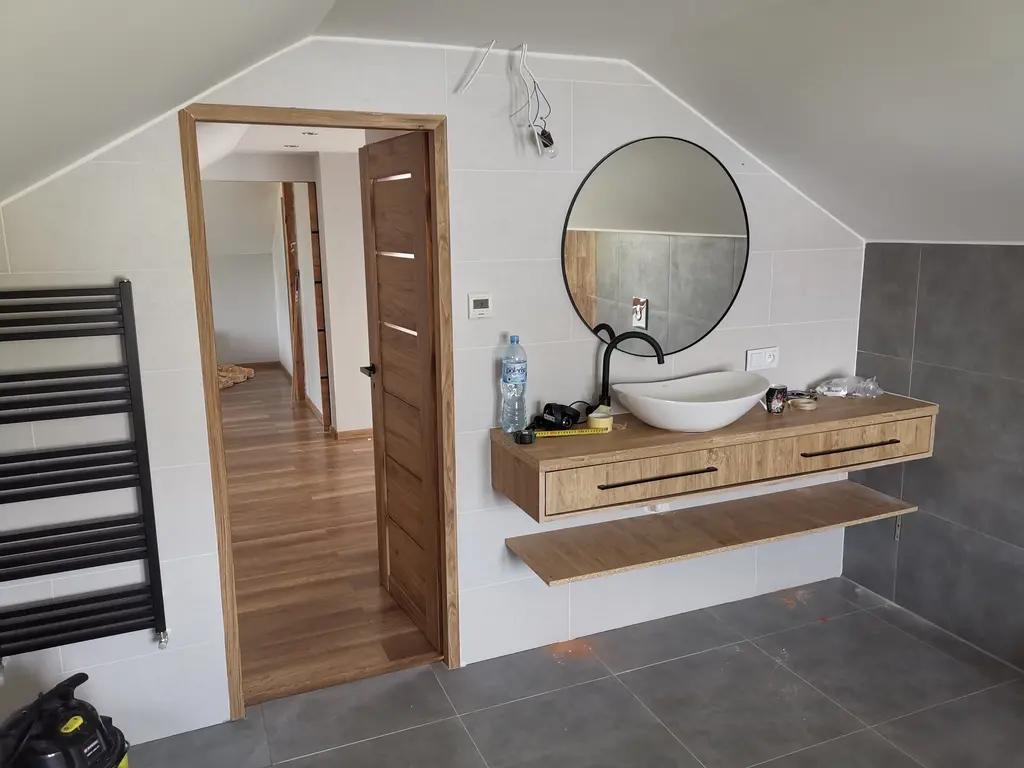 Nowoczesna łazienka na poddaszu z drewnianą szafką pod umywalką, okrągłym lustrem i otwartymi drzwiami ukazującymi parkiet. Widoczny grzejnik drabinkowy.