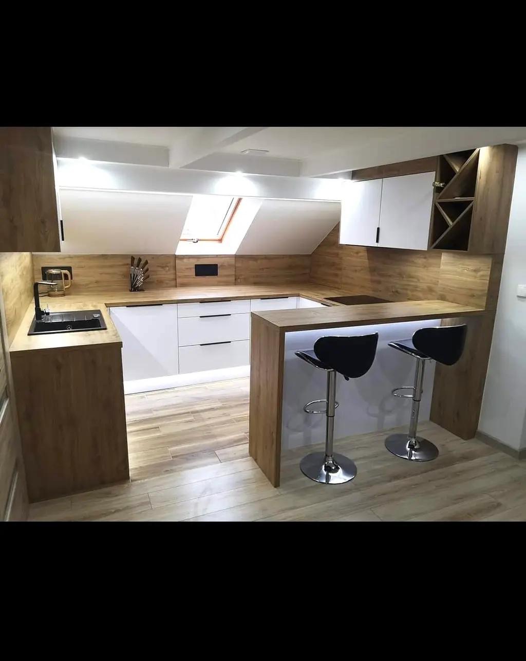 Nowoczesna kuchnia w skosach: białe fronty, drewniane blaty i barkiem z hokerami. Uwagę przykuwa skośne okno dachowe i oświetlenie LED pod blatem.