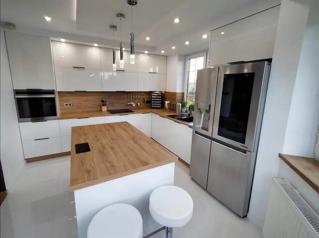 Nowoczesna kuchnia z białymi, lakierowanymi frontami, drewnianym blatem i wyspą. Widoczne srebrna lodówka i dwa białe hokery. Oświetlenie punktowe i wiszące lampy.