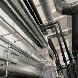 Anter Anna Pyrek - Różnorodne rury instalacyjne (czarne, srebrne, izolowane) prowadzone pod sufitem w budynku. Widoczne wsporniki i elementy mocujące. Ujęcie z dołu, perspektywa podkreśla złożoność instalacji.