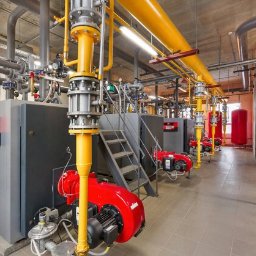 Anter Anna Pyrek - Kotłownia z rzędami pieców gazowych, żółtymi rurami i szarą instalacją. Widoczne schody i czerwone elementy pieców. Ujęcie z perspektywy podłogi.