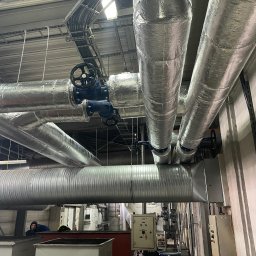 Anter Anna Pyrek - Izolowane rury grzewcze z zaworami w hali przemysłowej, widok z dołu. Instalacja z elementami sterującymi i pracownikiem w tle. Metalowe rury z niebieskimi zaworami.