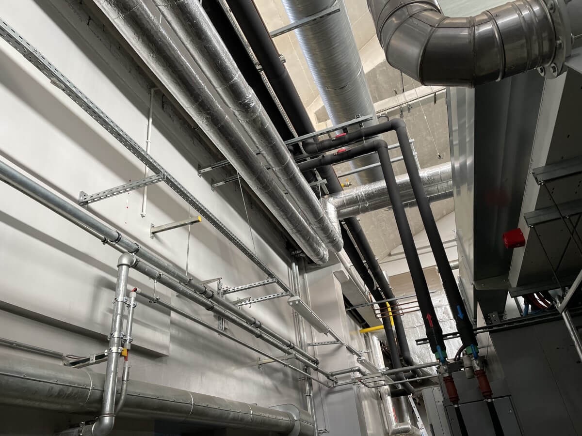Różnorodne rury instalacyjne (czarne, srebrne, izolowane) prowadzone pod sufitem w budynku. Widoczne wsporniki i elementy mocujące. Ujęcie z dołu, perspektywa podkreśla złożoność instalacji.