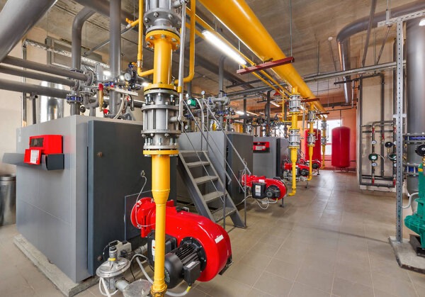 Kotłownia z rzędami pieców gazowych, żółtymi rurami i szarą instalacją. Widoczne schody i czerwone elementy pieców. Ujęcie z perspektywy podłogi.