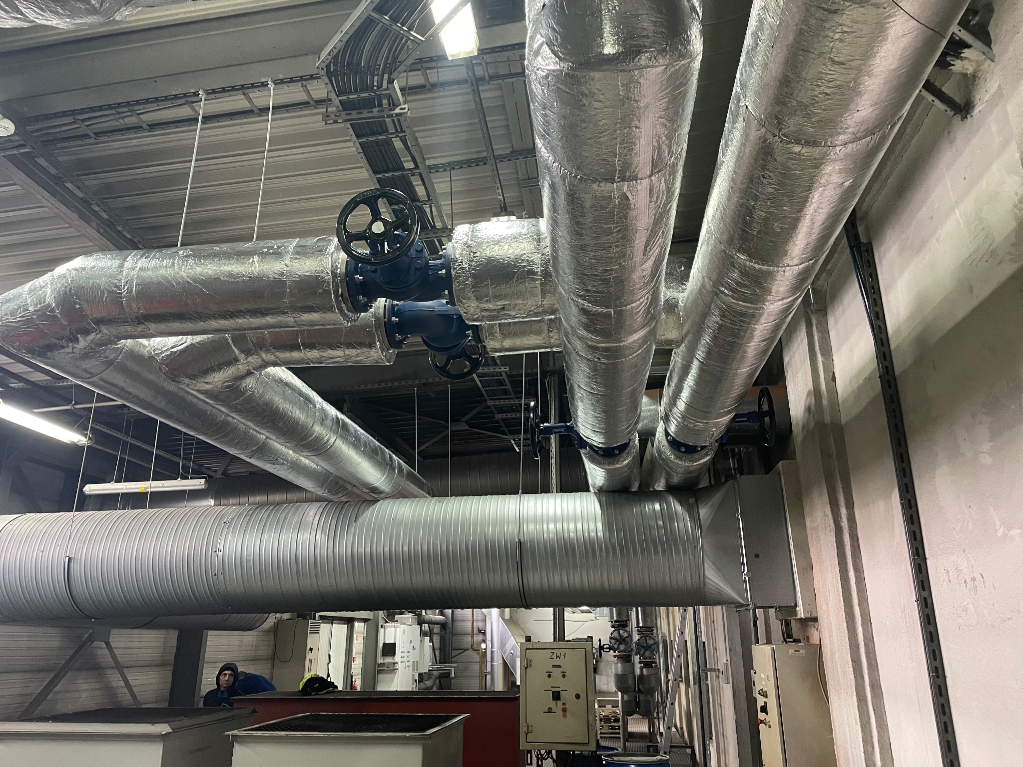 Izolowane rury grzewcze z zaworami w hali przemysłowej, widok z dołu. Instalacja z elementami sterującymi i pracownikiem w tle. Metalowe rury z niebieskimi zaworami.