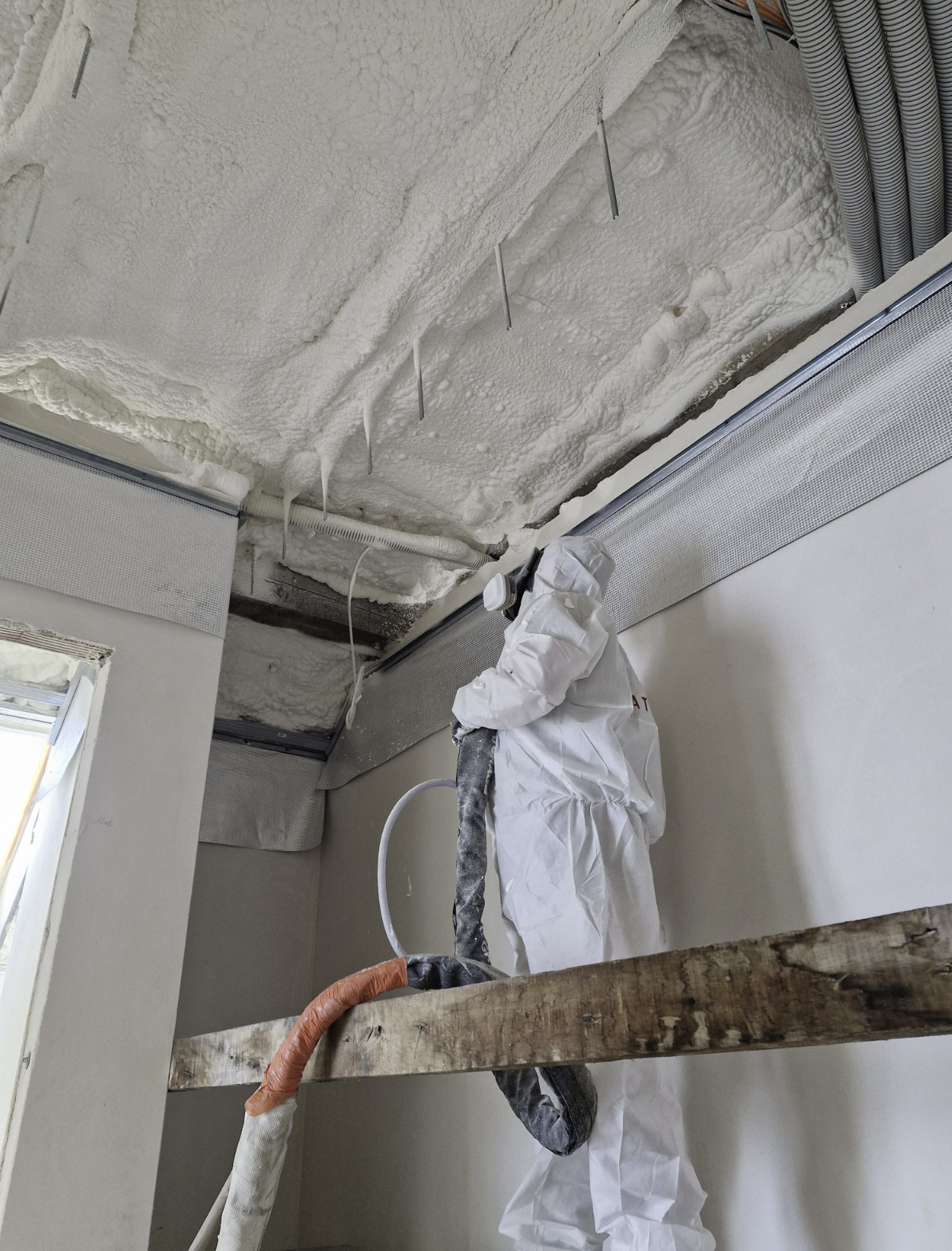 Pracownik w kombinezonie ochronnym aplikuje pianę izolacyjną na sufit wewnątrz budynku. Widoczne elementy konstrukcyjne i przewody.