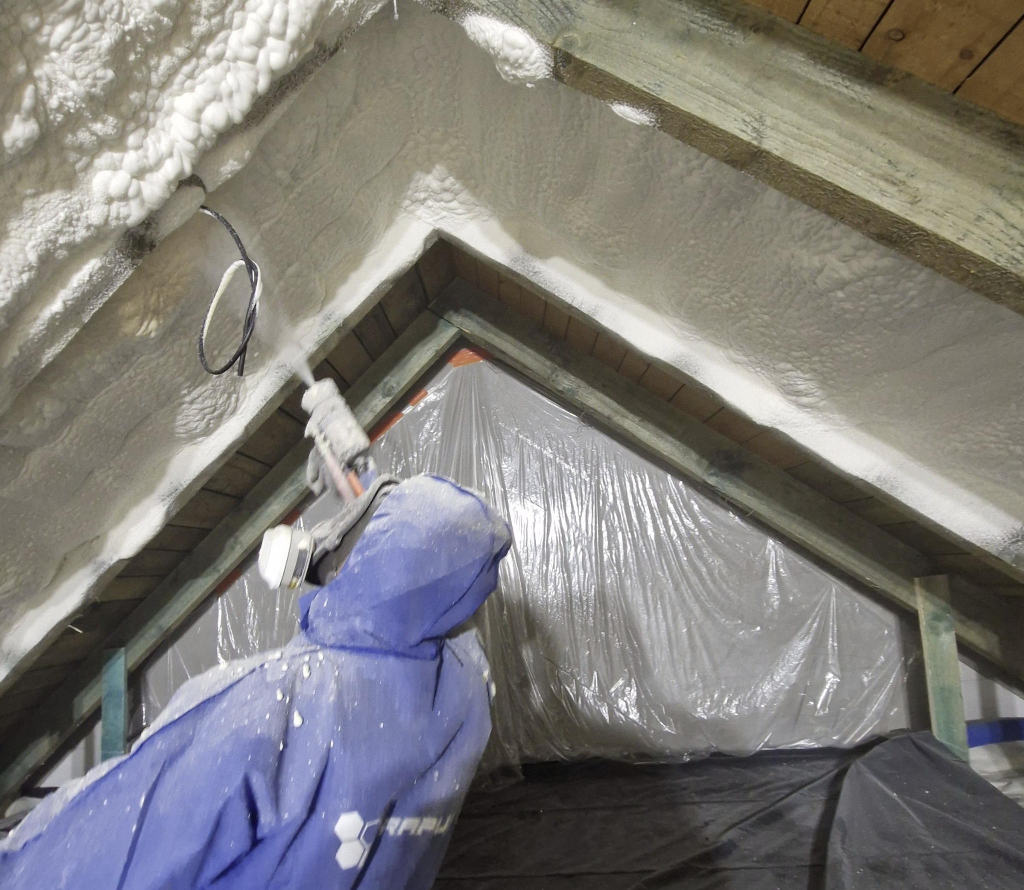 Aplikacja izolacji piankowej na poddaszu z widocznym pracownikiem w kombinezonie ochronnym i narzędziem do natrysku, zabezpieczona folią. Detal wykończenia.