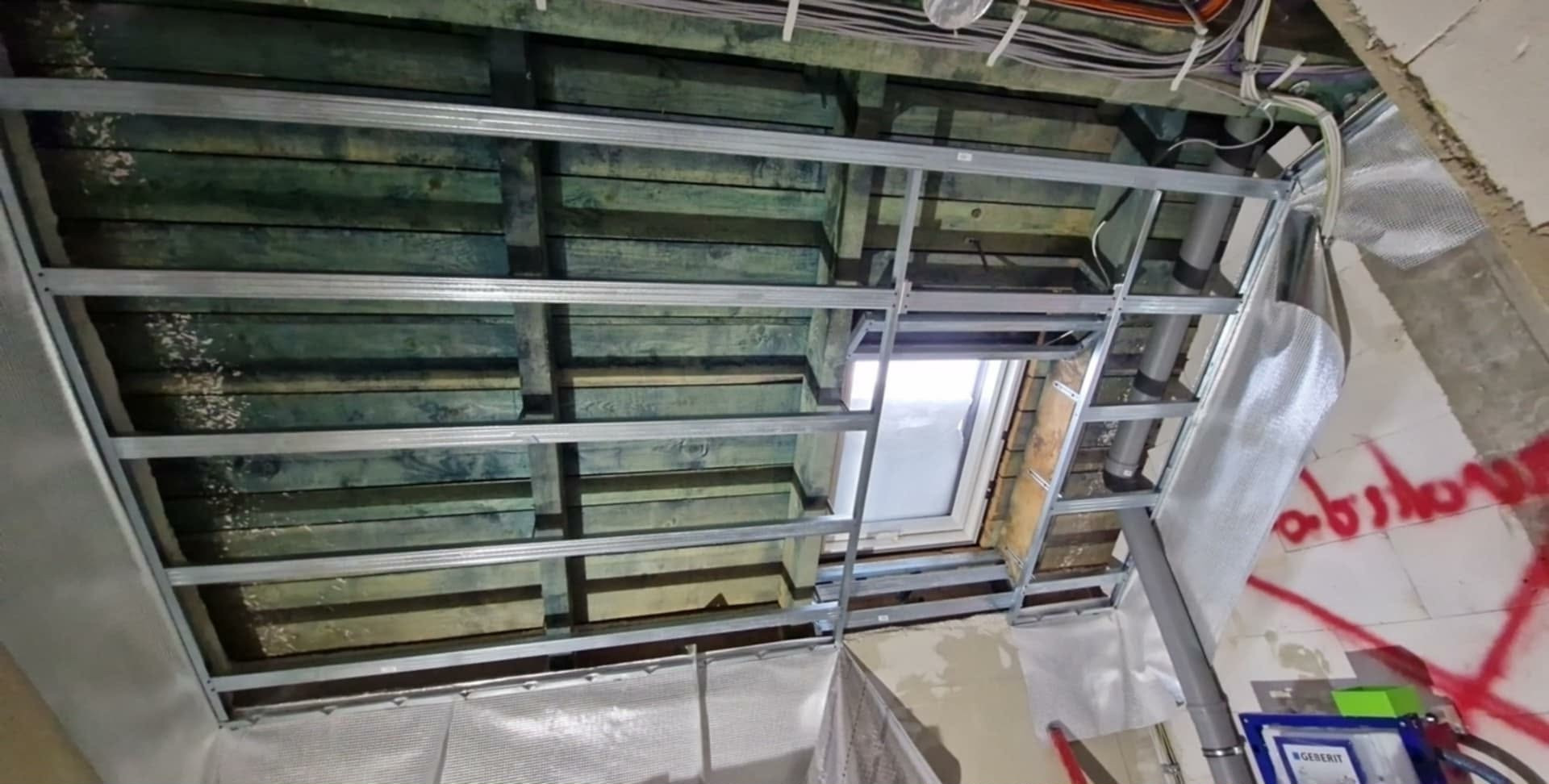 Sufit z drewnianą konstrukcją i metalowym stelażem pod płyty gipsowe, widoczne przewody i rury, okno dachowe. Etap prac wykończeniowych.