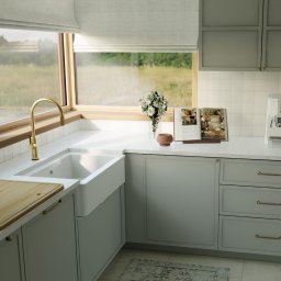 Materna Design - Jasna kuchnia z oknem na zielony krajobraz. Biały zlew farmerski, złota bateria, drewniana deska do krojenia. Stonowana kolorystyka szafek i blatu. Ekspres do kawy.