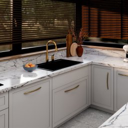 Materna Design - Elegancka kuchnia z marmurowym blatem, złotą armaturą i białymi szafkami. Widoczne detale: ekspres do kawy Smeg, owoce w misce i drewniane żaluzje.