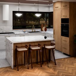 Materna Design - Nowoczesna kuchnia z marmurowym blatem wyspy i drewnianymi szafkami. Parkiet w jodełkę, minimalistyczne lampy i rolety w oknach tworzą elegancki wystrój.