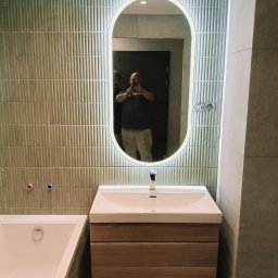 Marceli Wilaszek - Nowoczesna łazienka w Chorzowie po remoncie: zielone płytki na ścianie, owalne lustro z podświetleniem LED, umywalka z drewnianą szafką i wanna. Widoczne instalacje.