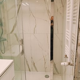 Marceli Wilaszek - Nowoczesna kabina prysznicowa z białymi płytkami imitującymi marmur i grzejnikiem drabinkowym. Widoczny fragment szafki umywalkowej.