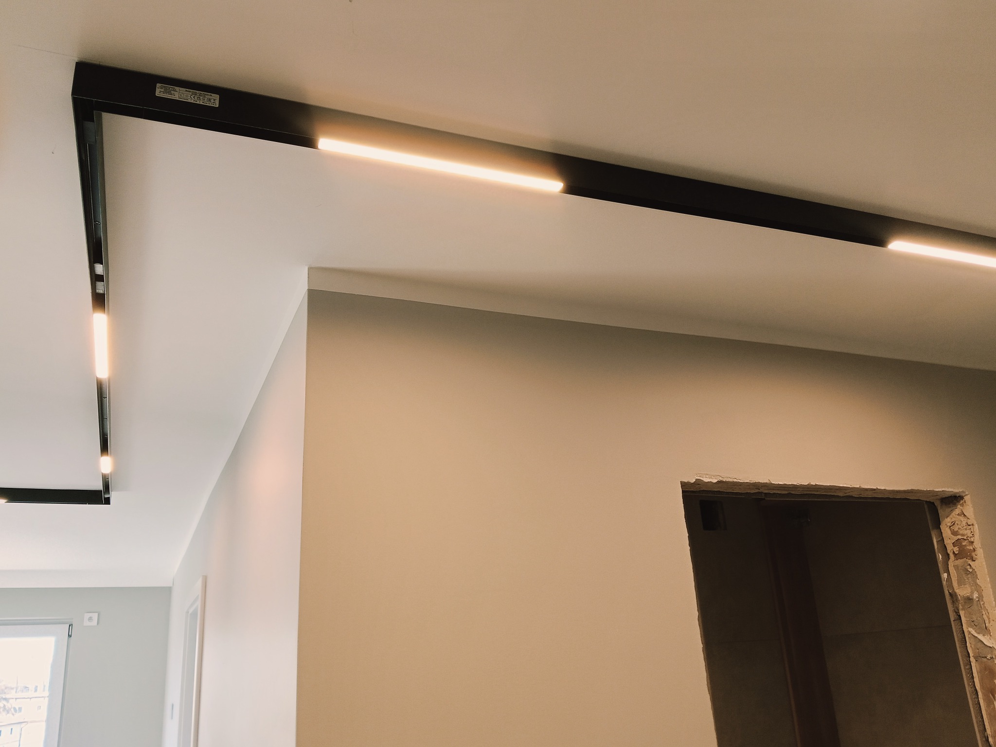 Nowoczesne oświetlenie sufitowe z czarnymi szynami i liniowymi lampami LED w jasnym wnętrzu. Widoczny fragment ściany i otwór drzwiowy w stanie surowym.