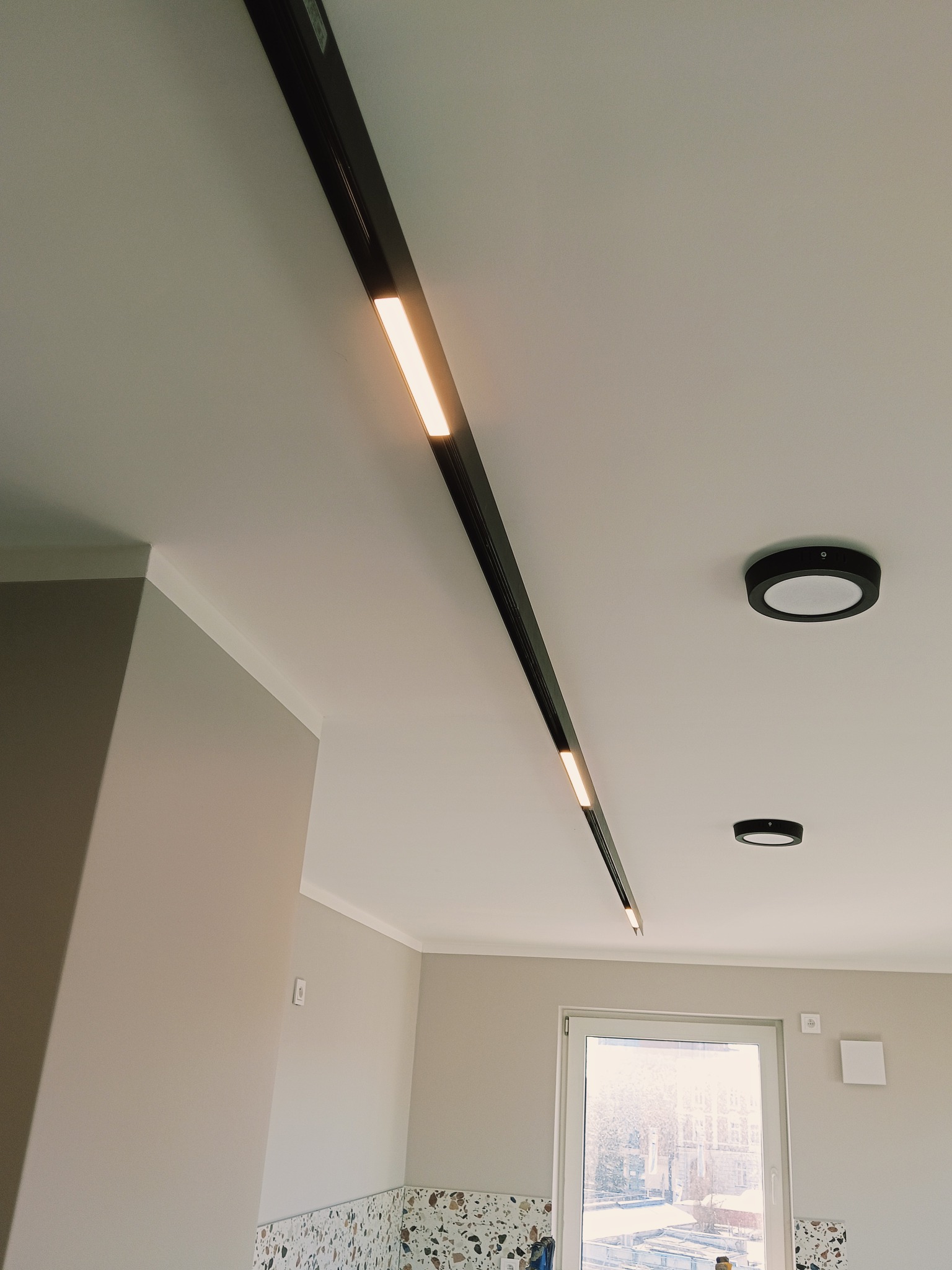 Nowoczesne oświetlenie sufitowe: czarna szyna z wbudowanymi lampami LED, uzupełniona okrągłymi lampami natynkowymi. Widok na jasne wnętrze z oknem i fragmentem ściany.