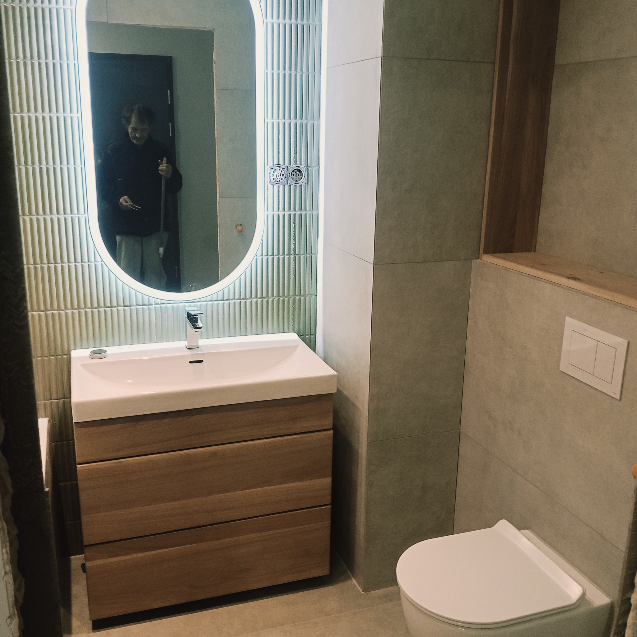 Nowoczesna łazienka z umywalką na drewnianej szafce, podświetlanym lustrem i toaletą. Ściany wyłożone płytkami w odcieniach szarości i zieleni. W lustrze odbija się osoba.