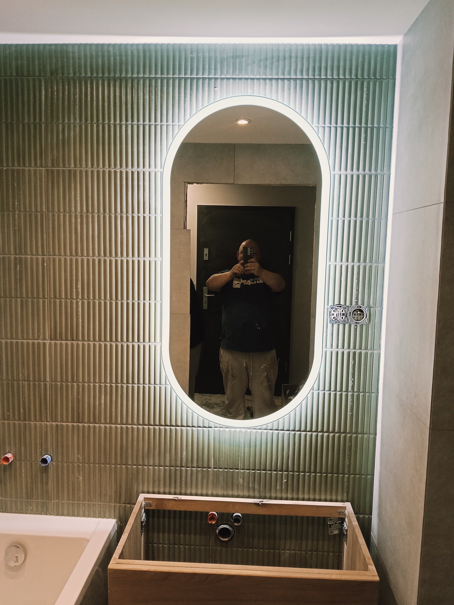 Montaż owalnego lustra z podświetleniem LED na ścianie z zielonych, żłobionych płytek w łazience. Widoczna niedokończona szafka pod umywalkę i odbicie osoby robiącej zdjęcie.