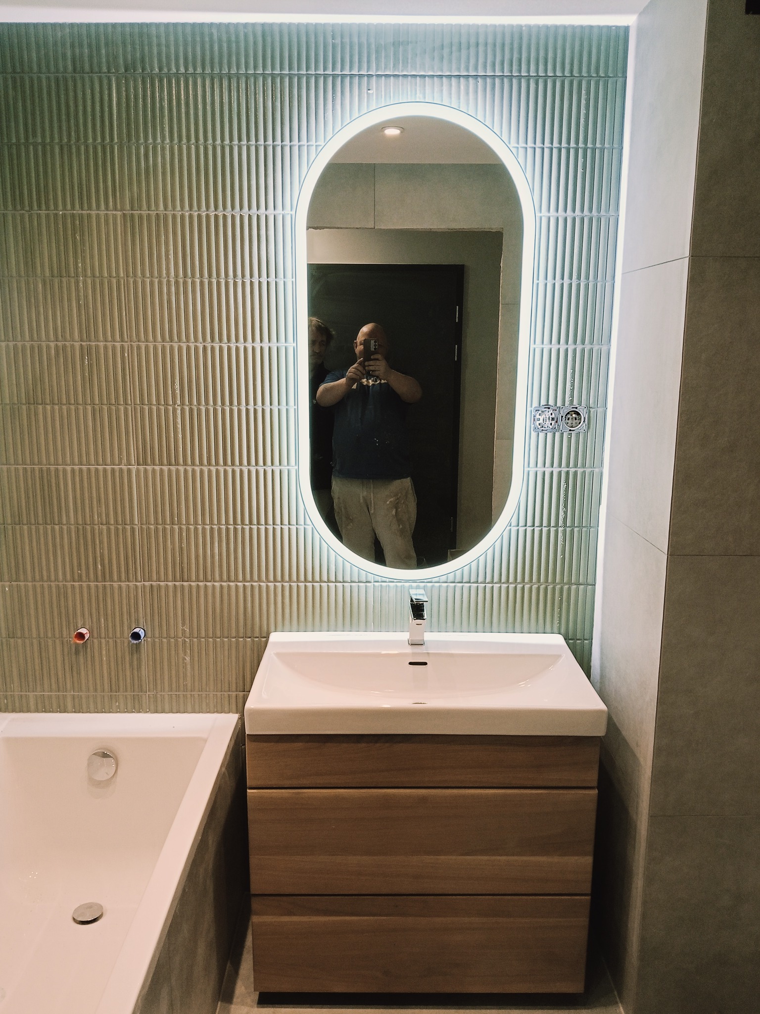 Nowoczesna łazienka w Chorzowie po remoncie: zielone płytki na ścianie, owalne lustro z podświetleniem LED, umywalka z drewnianą szafką i wanna. Widoczne instalacje.