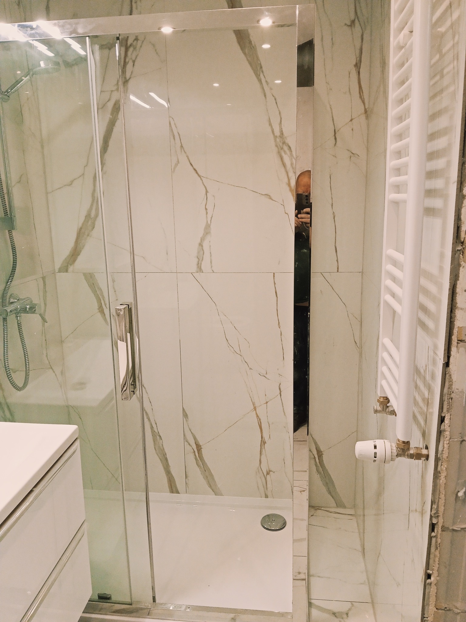 Nowoczesna kabina prysznicowa z białymi płytkami imitującymi marmur i grzejnikiem drabinkowym. Widoczny fragment szafki umywalkowej.