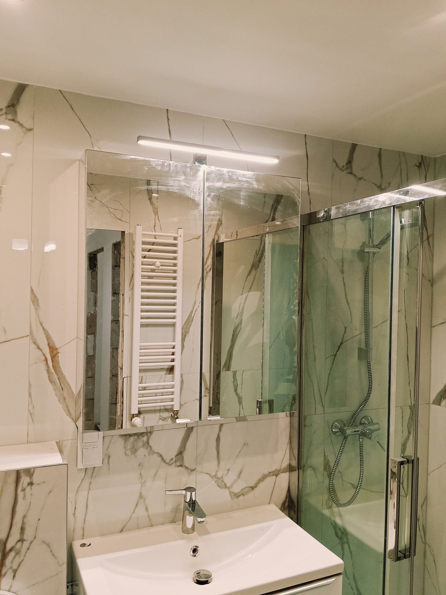 Wykończona łazienka z kabiną prysznicową i umywalką, marmurowe płytki na ścianach, lustro z oświetleniem. Widoczny grzejnik drabinkowy.