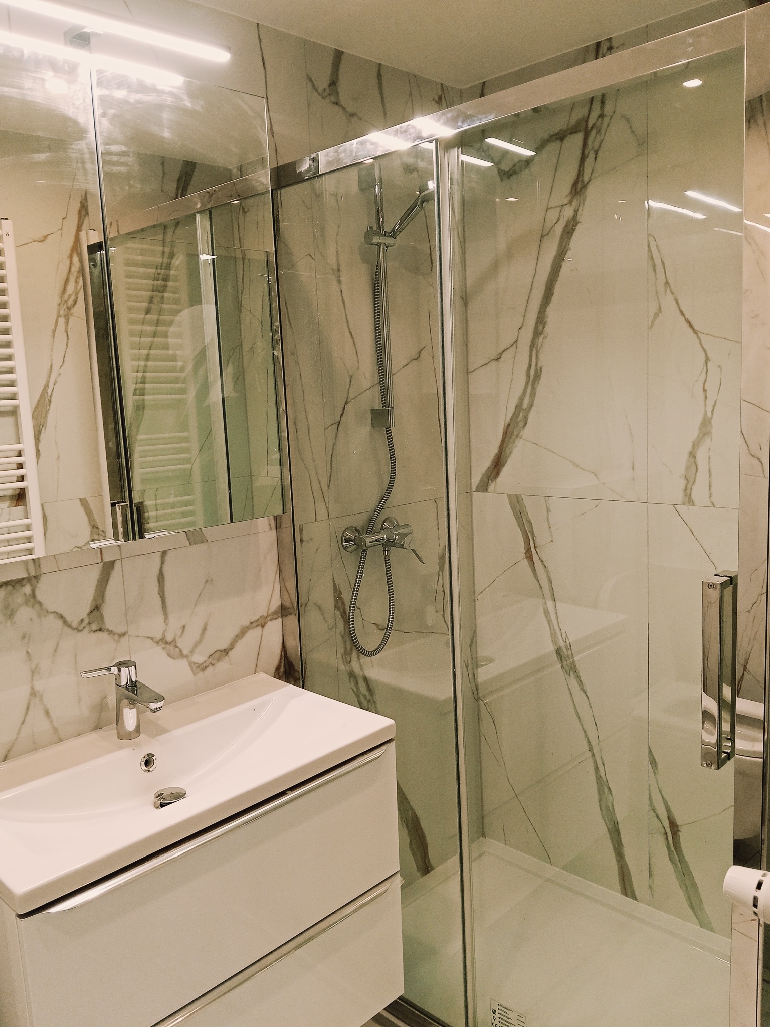 Nowoczesna łazienka z kabiną prysznicową i umywalką w szafce, wykończona białymi płytkami imitującymi marmur z wyraźnym, szarym żyłowaniem. Chromowana armatura.