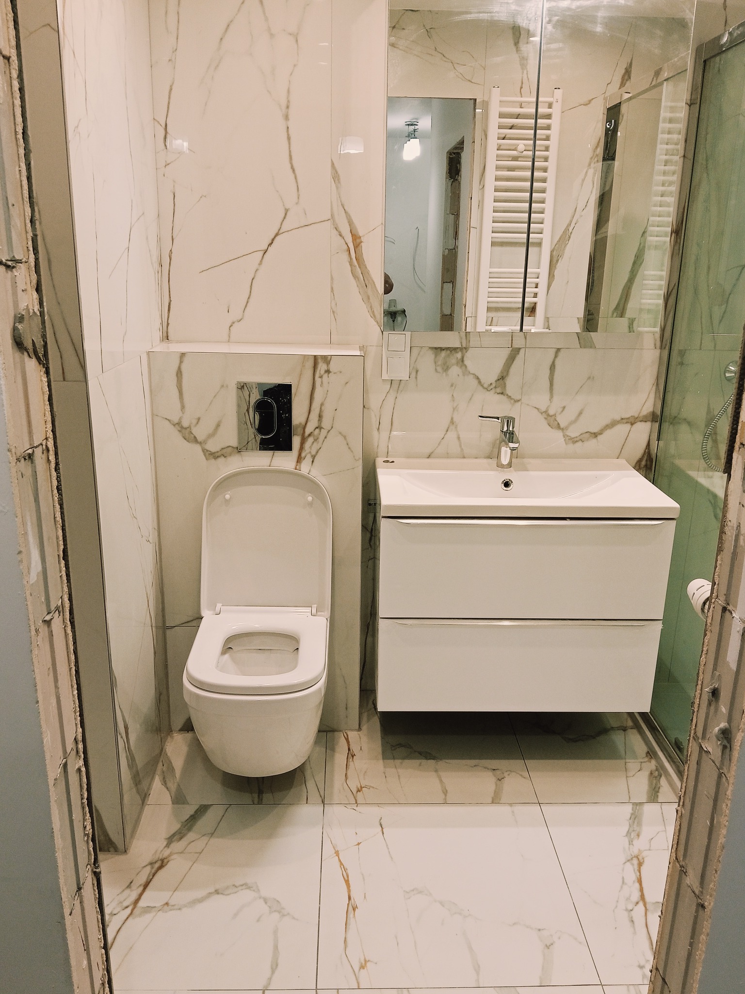 Wykończona łazienka z białymi płytkami imitującymi marmur, wisząca toaleta i szafka z umywalką. Widoczne fragmenty niedokończonego wykończenia przy futrynie.