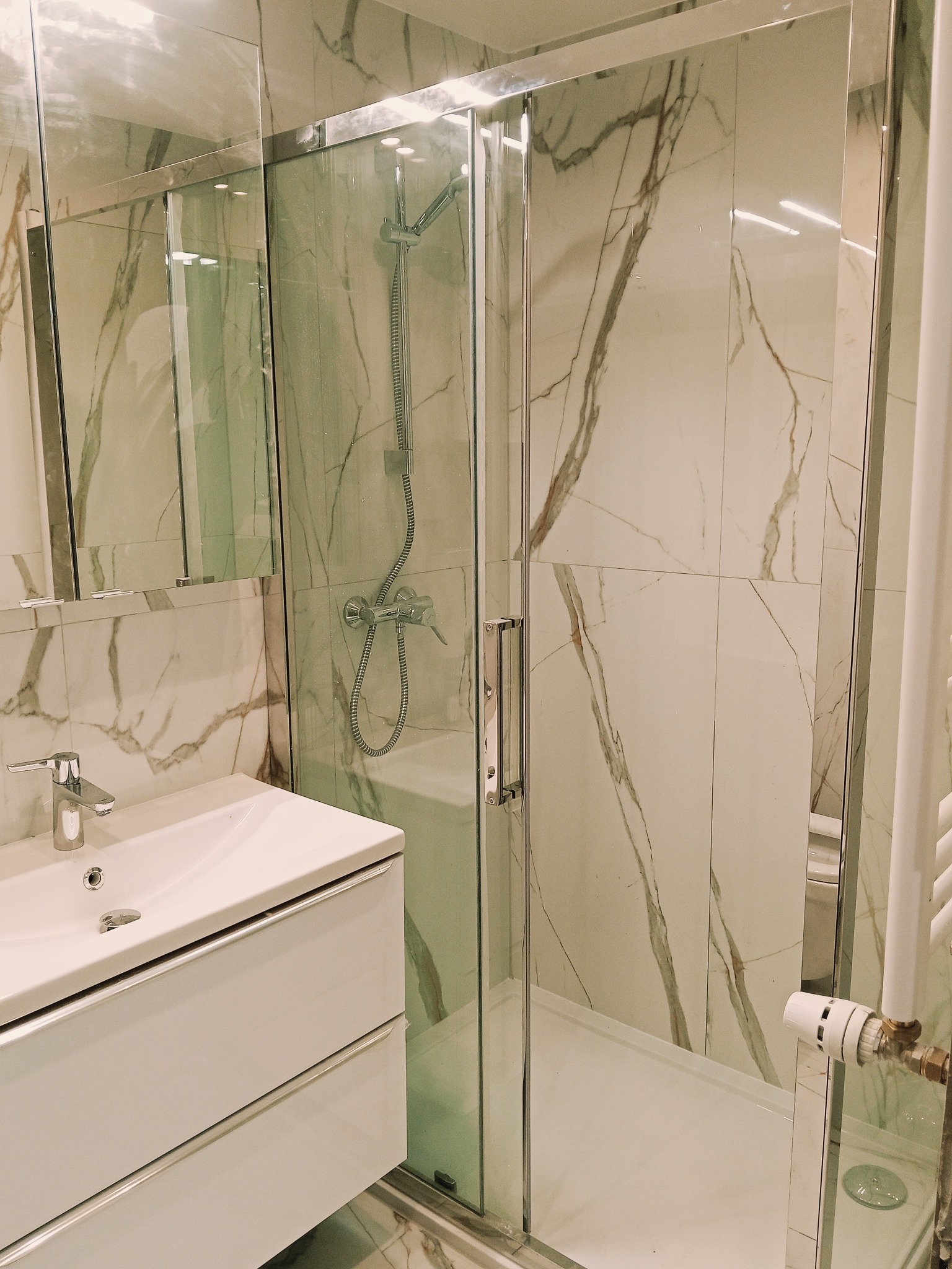 Nowoczesna łazienka z kabiną prysznicową typu walk-in, białą umywalką z szafką i marmurowymi płytkami na ścianach. Chromowane wykończenia i minimalistyczny design.