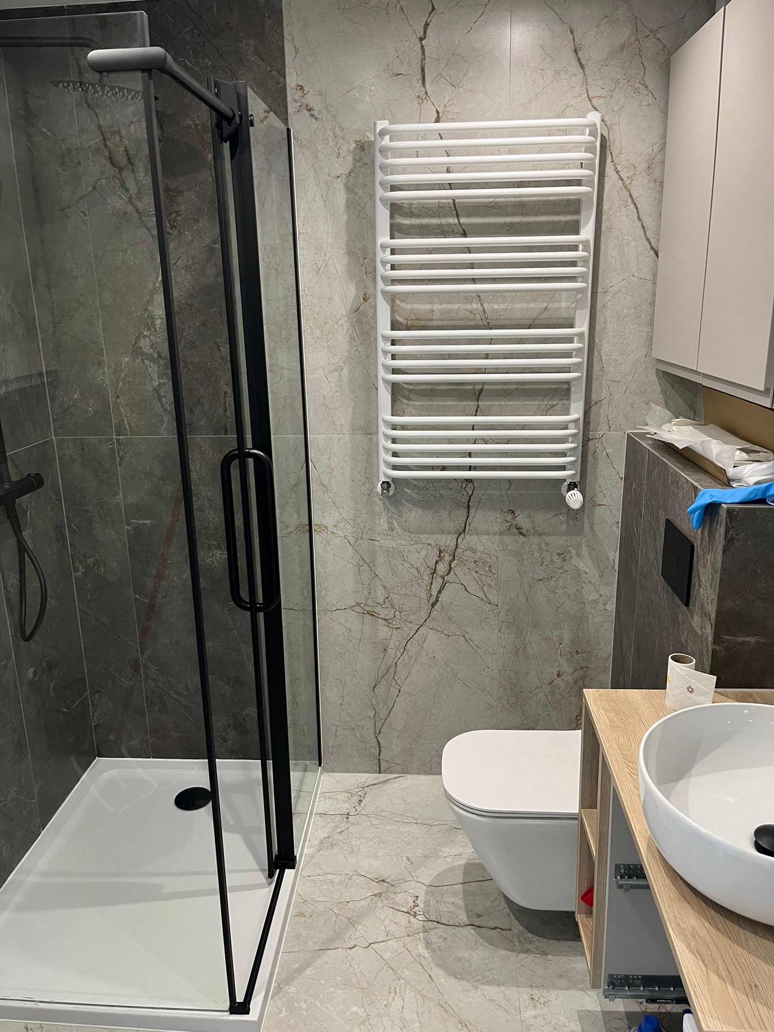 Nowoczesna łazienka po remoncie: kabina prysznicowa z czarnymi ramami, biały grzejnik drabinkowy i umywalka na drewnianej szafce. Marmurowe płytki na ścianach i podłodze.