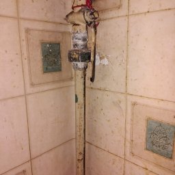 Wolkgaz24na7 - Stara, pionowa rura gazowa z zaworem, zabezpieczona czerwoną taśmą, na tle ściany wyłożonej kwadratowymi, beżowymi płytkami z dekoracyjnym motywem.