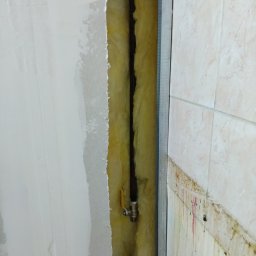 Wolkgaz24na7 - Odsłonięta instalacja gazowa w ścianie z izolacją, widoczny zawór. Fragment ściany z płytkami i druga ściana w trakcie wykończenia. Metalowy profil.