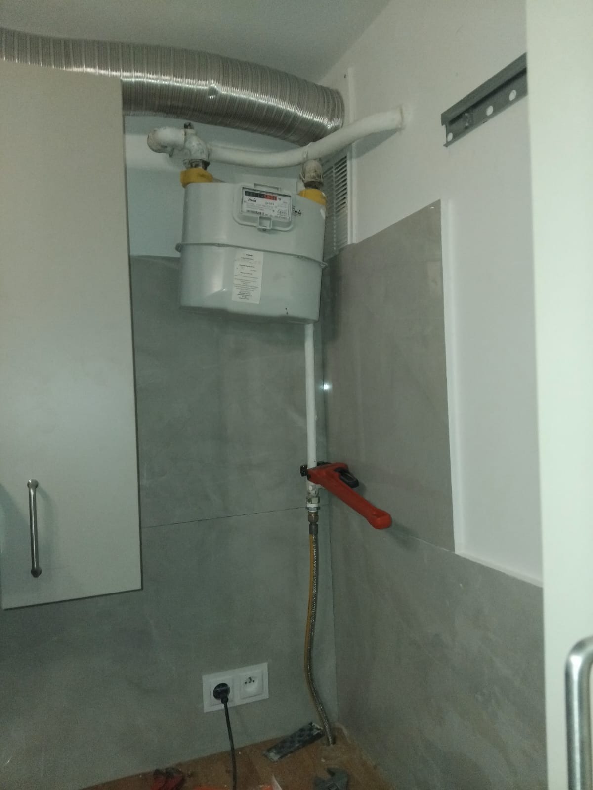 Szary gazomierz z rurami w jasnym pomieszczeniu, widoczny klucz nastawny i gniazdko elektryczne. Element instalacji gazowej w trakcie prac.