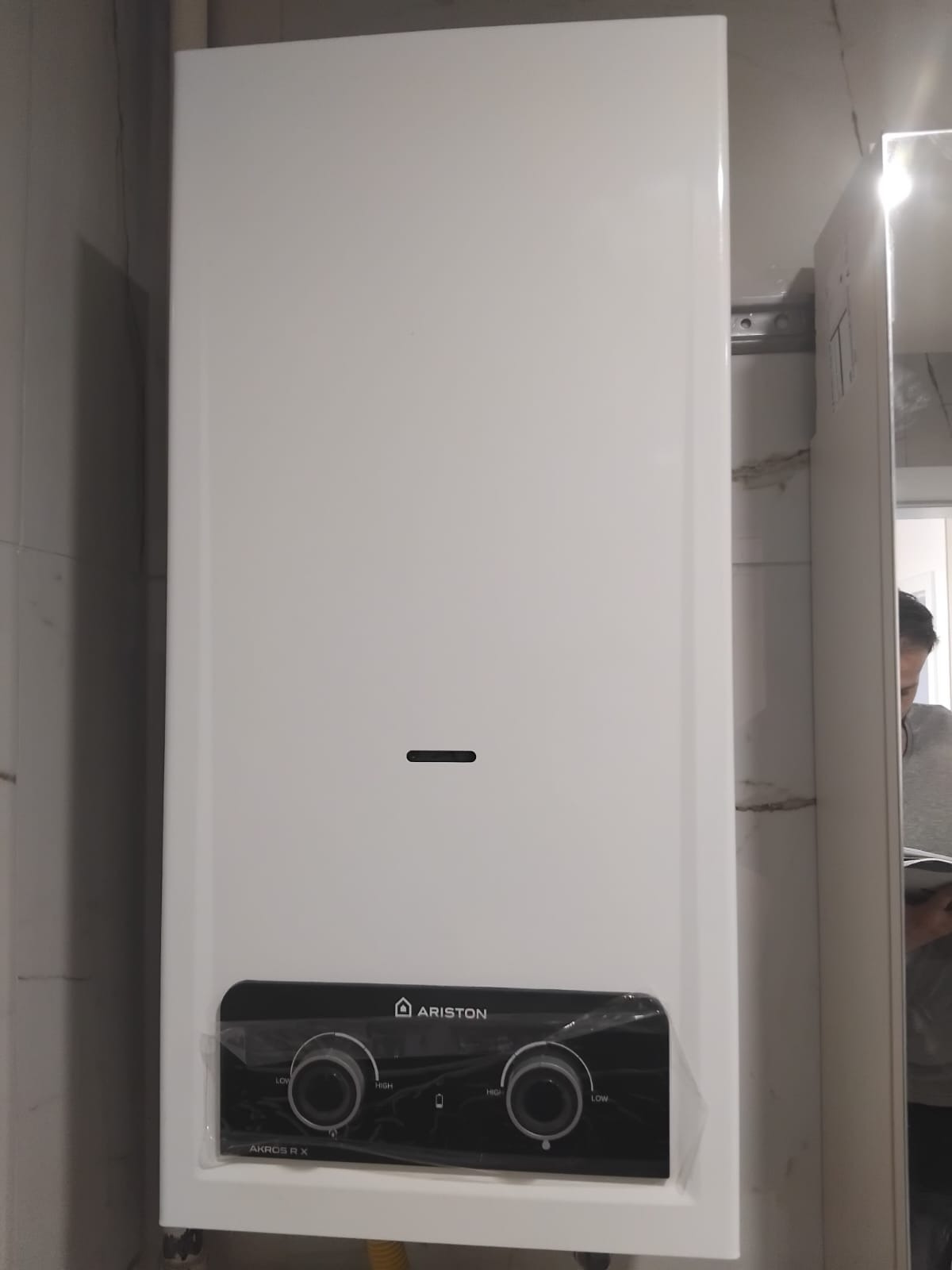 Biały, wiszący piec gazowy Ariston Akros RX z czarnym panelem sterowania, jeszcze w folii ochronnej, zamontowany na ścianie z jasnymi płytkami. Widoczny fragment żółtego węża.