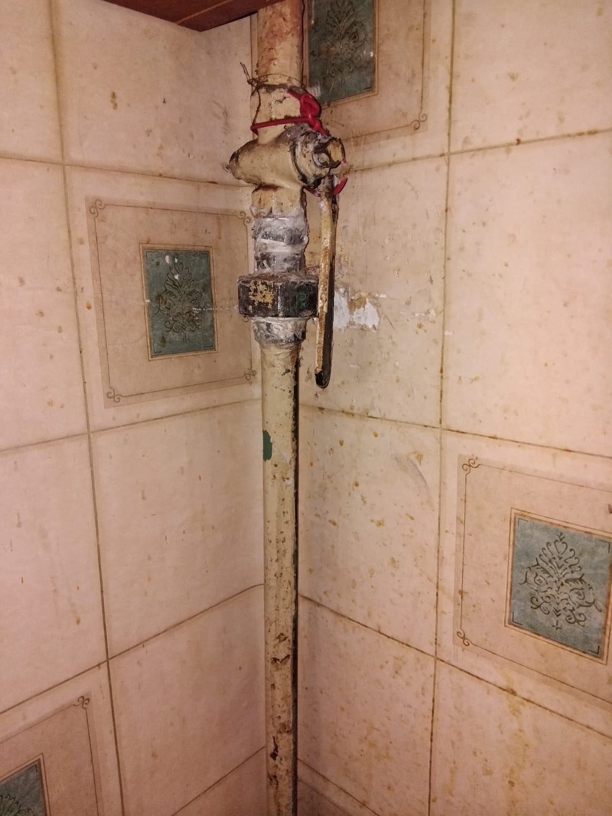 Stara, pionowa rura gazowa z zaworem, zabezpieczona czerwoną taśmą, na tle ściany wyłożonej kwadratowymi, beżowymi płytkami z dekoracyjnym motywem.