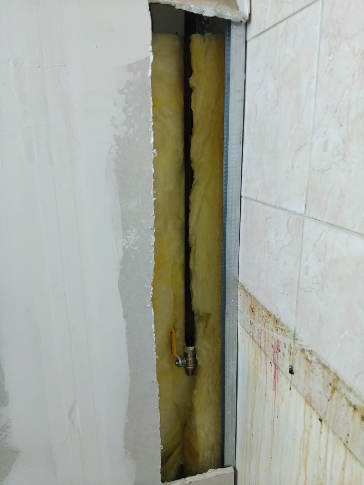 Odsłonięta instalacja gazowa w ścianie z izolacją, widoczny zawór. Fragment ściany z płytkami i druga ściana w trakcie wykończenia. Metalowy profil.